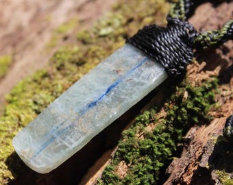 May include: A pendant featuring a long, rectangular, translucent light blue kyanite crystal with a distinct dark blue streak running down its center, securely wrapped at the top with black cord that transitions into a braided green and black necklace.