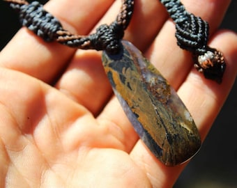 May include: A pendant necklace featuring an elongated, polished stone with swirling patterns of brown, black, and translucent purple. The stone is suspended from a dark brown and black macrame cord with intricate knotted details and a small silver bead.