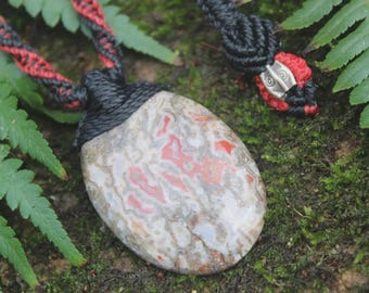May include: A polished, oval-shaped stone pendant featuring a mottled pattern of red, white, and grey tones. The stone is secured by a black and red macramé cord, which includes a silver-toned metal bead accent near the adjustable closure.