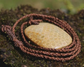 May include: A smooth, oval-shaped fossilized coral pendant with a tan and beige speckled pattern. The stone is suspended on a dark brown, braided cord necklace that is coiled around the pendant, featuring a knotted adjustable sliding closure.