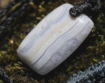 May include: A rectangular blue lace agate pendant with soft white and light blue banding, featuring a smooth, polished surface. The stone is suspended from a thick, braided black cord that loops through a hole at the top of the gemstone.