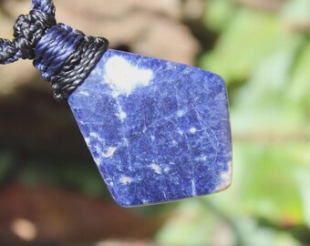 May include: A pendant made of a polished, irregular pentagonal piece of deep blue sodalite stone with white mineral inclusions. The stone is secured by a dark blue and black braided cord that wraps around the top edge of the pendant.