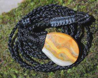 May include: A teardrop-shaped bumblebee jasper pendant with yellow, orange, and white bands, secured by a black braided cord necklace featuring intricate macrame knotting and an adjustable sliding closure mechanism.