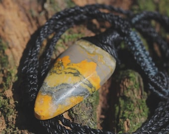 May include: A polished, teardrop-shaped pendant featuring a marbled pattern of vibrant yellow, charcoal gray, and white tones. The stone is securely attached to a thick, braided black cord necklace that loops around the top of the pendant.