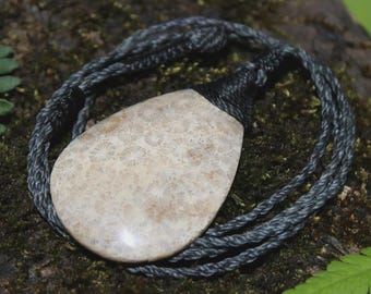 May include: A teardrop-shaped pendant made of fossilized coral with a beige and tan starburst pattern. The pendant is suspended from a dark gray, multi-strand twisted cord that is knotted securely at the top of the stone setting.