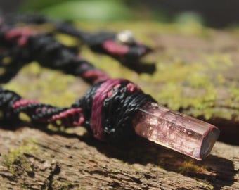 May include: A necklace featuring a raw, translucent pink crystal pendant secured by black and dark red macrame cord. The cord is intricately knotted in a spiral pattern, holding the elongated, faceted gemstone firmly in place at the center.