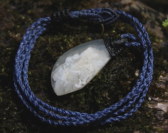 May include: A pendant necklace featuring a translucent, teardrop-shaped stone with white, cloud-like inclusions. The stone is secured by a black cord wrap and suspended from a multi-strand, braided dark blue cord with adjustable sliding knots.