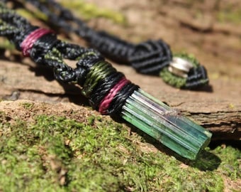 May include: A pendant featuring a long, translucent green and pink tourmaline crystal secured by black, olive green, and magenta cord macrame. The cord is intricately knotted with a silver-toned bead accent near the adjustable closure mechanism.