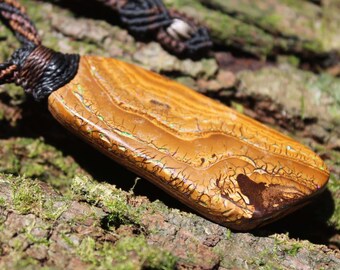 May include: A rectangular boulder opal pendant featuring a polished tan and brown surface with intricate, dark, vein-like inclusions and subtle iridescent green flashes. The stone is secured by a black and brown braided cord necklace.