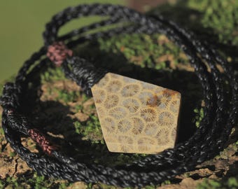 May include: A pendant necklace featuring a polished, pentagonal-shaped piece of fossilized coral with a tan and brown starburst pattern. The stone is suspended from a black braided cord with dark copper-colored accents at the adjustable knots.