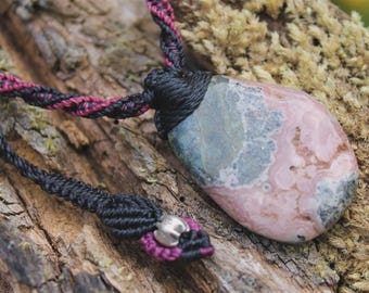 May include: A pendant necklace featuring a smooth, teardrop-shaped stone with mottled pink, gray, and white patterns. The stone is secured by a black and magenta macramé cord, which is intricately knotted and includes a small silver-toned metal bead.