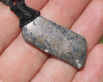 May include: A rectangular pendant featuring a mottled pattern of grey, black, and metallic gold tones, suspended from a thick black braided cord. The stone surface displays an intricate, organic texture with varied mineral inclusions throughout.