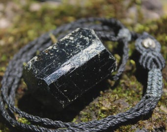 May include: A raw, black tourmaline crystal pendant secured by a dark gray, braided cord necklace. The stone features a natural, hexagonal prismatic structure with a glossy, reflective surface and visible striations along its vertical length.