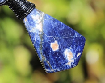 May include: A polished, irregularly shaped blue sodalite pendant featuring white and tan mineral inclusions, suspended from a thick black cord. The stone displays a deep royal blue base color with visible veining and distinct light-colored patches.