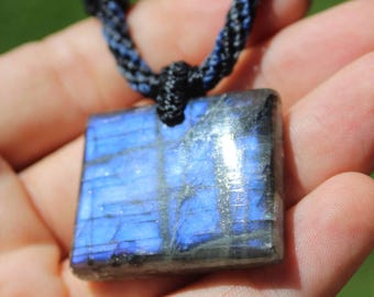 May include: A square labradorite pendant featuring a vibrant blue iridescent flash and dark grey inclusions. The stone is suspended from a thick, braided black cord necklace that attaches to the top of the pendant with a secure knot.