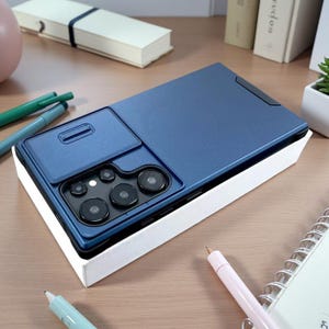 Puede incluir: Una funda de smartphone azul oscuro con una cubierta deslizante para el objetivo de la cámara. La funda tiene un recorte rectangular para el conjunto de cámaras, que incluye cinco lentes circulares y un flash, protegidos por la cubierta.