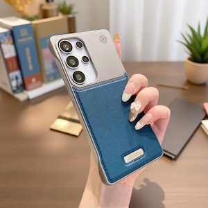 May include: A smartphone case featuring a two-tone design with a silver-colored upper section containing a circular speaker grille and a textured blue lower section. A small rectangular silver metal plate is attached to the bottom of the blue area.