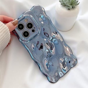 Puede incluir: Una funda de teléfono azul con una superficie reflectante similar a un líquido y un borde irregular y ondulado. La funda presenta un recorte cuadrado para la cámara con tres aberturas circulares para lentes y un flash para smartphone.