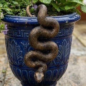 Puede incluir: Una escultura de serpiente color bronce con una superficie texturizada y escamosa enroscada sobre el borde de una maceta de cerámica azul profundo. La maceta presenta un patrón de greca griega en la parte superior y motivos de hojas en relieve.