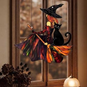 Pode incluir: Um apanhador de sol em vitral representando uma figura com um chapéu pontiagudo preto e um vestido fluido em tons de laranja, vermelho e roxo, montada numa vassoura. Um gato preto sólido está sentado na vassoura ao lado da figura, suspensos por fios.