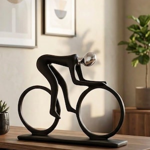 Può includere: Una scultura nero opaco di un ciclista in posizione da corsa, caratterizzata da una testa argentata liscia e riflettente. La figura è montata su una base rettangolare nera, con due ruote circolari integrate nel telaio della bicicletta.