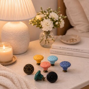 Puede incluir: Una colección de siete pomos de cerámica redondos para armarios con intrincados patrones florales en relieve. Los pomos están acabados en varios colores, incluyendo amarillo, azul claro, coral, azul oscuro, negro y verde, cada uno con una base metálica.