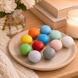 Puede incluir: Una colección de doce pomos de armario redondos y lisos en una variedad de colores sólidos, incluyendo rojo, naranja, amarillo, verde claro, azul, azul claro, verde menta, rosa, blanco, lavanda y gris, dispuestos sobre un plato de cerámica.