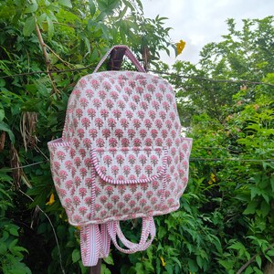 Op de afbeelding: Een gewatteerde rugzak met een herhalend patroon van kleine roze en groene bloemmotieven op een witte achtergrond. De tas heeft een voorvak met klep, twee zijvakken, een handvat aan de bovenkant en verstelbare schouderbanden met gestreepte rand.