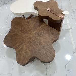 Könnte beinhalten: Ein Set aus drei ineinander verschachtelten Couchtischen mit organischen, amöbenartigen Formen. Ein Tisch hat eine glatte weiße Oberfläche, während die anderen beiden eine reiche braune Holzmaserung aufweisen. Ein kupferfarbener Streifen akzentuiert den kleinsten Tisch.