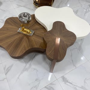 Könnte beinhalten: Ein Set aus drei ineinandergreifenden Couchtischen mit organischen, wolkenartigen Formen. Ein Tisch hat eine dunkle Holzmaserung, ein weiterer ist einfarbig weiß und der dritte ist ein kleineres dunkles Holzstück mit einem kupferfarbenen Bein.