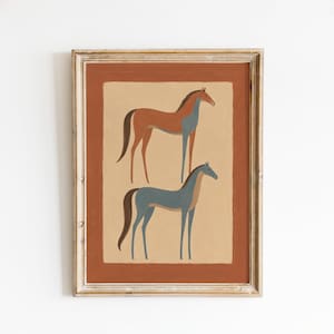 May include: A framed wall art print featuring two stylized horses standing one above the other. The top horse is brown with blue legs, and the bottom horse is blue with a brown tail. The background is a light tan rectangle set within a dark orange border.