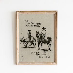 May include: A framed wall art print featuring a black ink illustration of two figures on horseback on a light beige background. The text reads: AND THE DREAMERS HAVE TOMORROW IF TODAY DOES NOT COME TRUE. The frame is a light-toned wood finish.