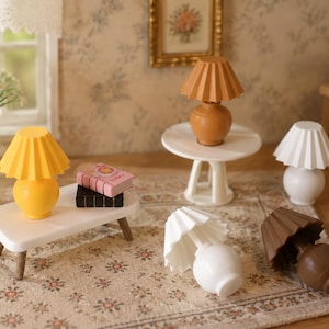 Puede incluir: Una colección de lámparas de mesa en miniatura con pantallas plisadas en amarillo, bronceado, blanco y marrón, junto a dos mesas blancas pequeñas. Una lámpara amarilla se sitúa sobre una mesa con dos libros diminutos al lado.