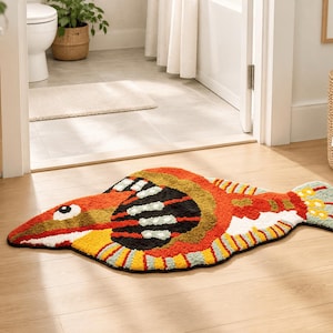 Puede incluir: Una alfombra de pelo cortado con forma de pez, que presenta una vibrante paleta de colores en naranja, amarillo, negro y verde claro. El diseño incluye un ojo prominente, escamas texturizadas y una aleta caudal bien definida.