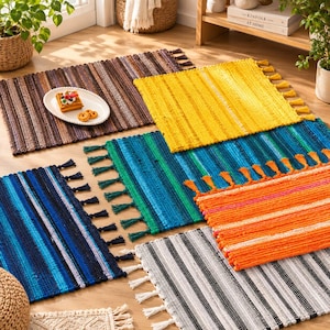 Puede incluir: Una colección de alfombras rectangulares tejidas con varios patrones de rayas en marrón, amarillo, azul, verde, naranja y gris. Cada alfombra está terminada con borlas decorativas en los bordes, mostrando un diseño de tela texturizada.