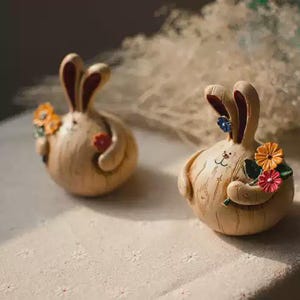 Puede incluir: Dos figuras de conejo redondas de color tostado claro con texturas de veta de madera. Cada conejo tiene orejas largas con interiores oscuros y sostiene un pequeño ramo de flores coloridas en tonos naranja, rojo y azul con ojos negros.