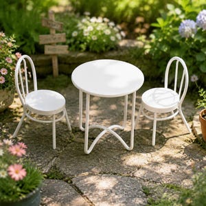 Puede incluir: Un conjunto de bistró en miniatura de color blanco que consta de una pequeña mesa redonda y dos sillas a juego con respaldos curvos. Los muebles presentan un acabado blanco sólido y liso con marcos de metal fino sobre un patio de piedra.