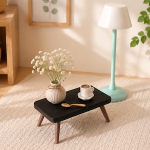 Puede incluir: Una mesa rectangular negra en miniatura con cuatro patas de madera, que presenta un pequeño jarrón blanco acanalado lleno de flores blancas de paniculata, una diminuta taza de café blanca sobre un platillo y una pequeña cuchara dorada.