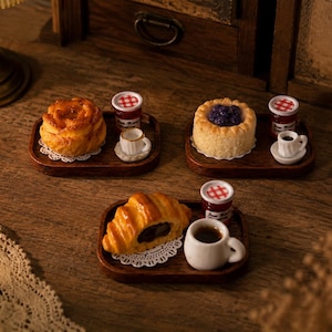 Puede incluir: Tres bandejas de madera en miniatura, cada una con un pequeño pastel, una diminuta taza de cerámica blanca con líquido oscuro y un pequeño frasco de vidrio con tapa de cuadros rojos y blancos. Los pasteles incluyen un bollo dorado, una tarta de arándanos y un cruasán de chocolate.