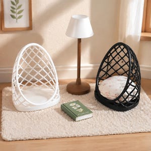 Puede incluir: Dos sillas en miniatura con forma de huevo y estructura de rejilla, una blanca y otra negra. La silla negra contiene un pequeño cojín redondo con estampado floral. Una lámpara de madera en miniatura con pantalla blanca y un pequeño libro verde titulado Flowers se sitúan entre ellas.