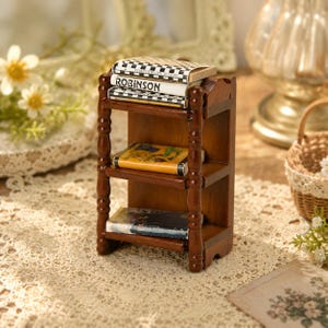 Puede incluir: Una estantería de madera en miniatura de tres niveles con acabado marrón oscuro y postes torneados. El estante superior contiene dos libros pequeños, uno con el lomo a cuadros blancos y negros y otro con la etiqueta ROBINSON. El estante central tiene un libro amarillo y el inferior un libro con cubierta azul oscuro.