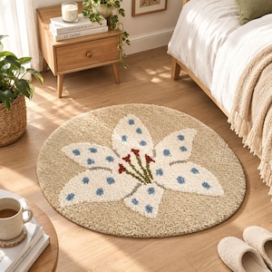 Op de afbeelding: Een rond, beige, getuft tapijt met een groot, wit, zesbladig leliemotief. De bloemblaadjes zijn versierd met kleine, lichtblauwe stippen en het midden van de bloem toont donkerrode en groene meeldraad-details.