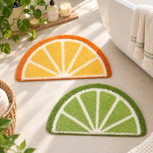 May include: Two semi-circular bath mats featuring a citrus fruit slice design. One mat is yellow and orange, resembling an orange slice, while the other is light and dark green, resembling a lime slice. Both mats have a textured, plush surface.
