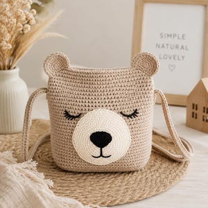 May include: A beige crochet crossbody bag shaped like a bear face with two small ears, closed eyes with eyelashes, a cream-colored muzzle, and a black embroidered nose and mouth. The bag features a matching beige crochet strap for carrying.