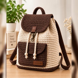 May include: A crochet backpack featuring a two-tone design with dark brown and cream yarn. The bag includes a top flap with a bronze-colored metal clasp, a front pocket with a brown leather-style tag labeled Handmade, and two shoulder straps.