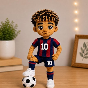 Puede incluir: Una muñeca de ganchillo hecha a mano de un jugador de fútbol con pelo castaño rizado, que lleva una camiseta y pantalones cortos a rayas azul marino y granate con el número 10 en blanco. La muñeca tiene un pie sobre un balón de fútbol de ganchillo.