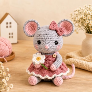 May include: A gray crocheted mouse toy with large pink-lined ears, wearing a dusty rose dress with a white scalloped hem and a small wooden button. The mouse has a pink nose, rosy cheeks, and holds a white daisy flower with a yellow center.
