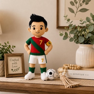 May include: A crocheted doll of a footballer with black hair, wearing a red and green jersey with the number 7, white shorts with the number 7, green socks, and white boots. The doll has a yellow captain's armband with a C and stands next to a football.