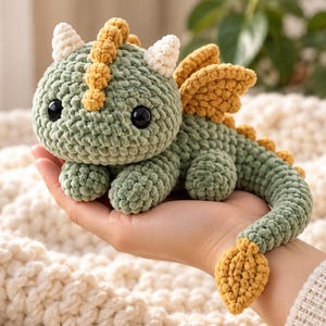 May include: A small, handmade crochet dragon plush toy featuring a sage green body, mustard yellow wings, and a matching yellow tail tip. The creature has two white horns, a yellow dorsal ridge, and large, round, black plastic safety eyes.