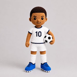 Puede incluir: Una muñeca de ganchillo hecha a mano de un jugador de fútbol con cabello castaño oscuro, vistiendo una camiseta blanca con el número 10 en azul oscuro, pantalones cortos blancos, calcetines blancos y botas azules. Sostiene un balón pequeño.